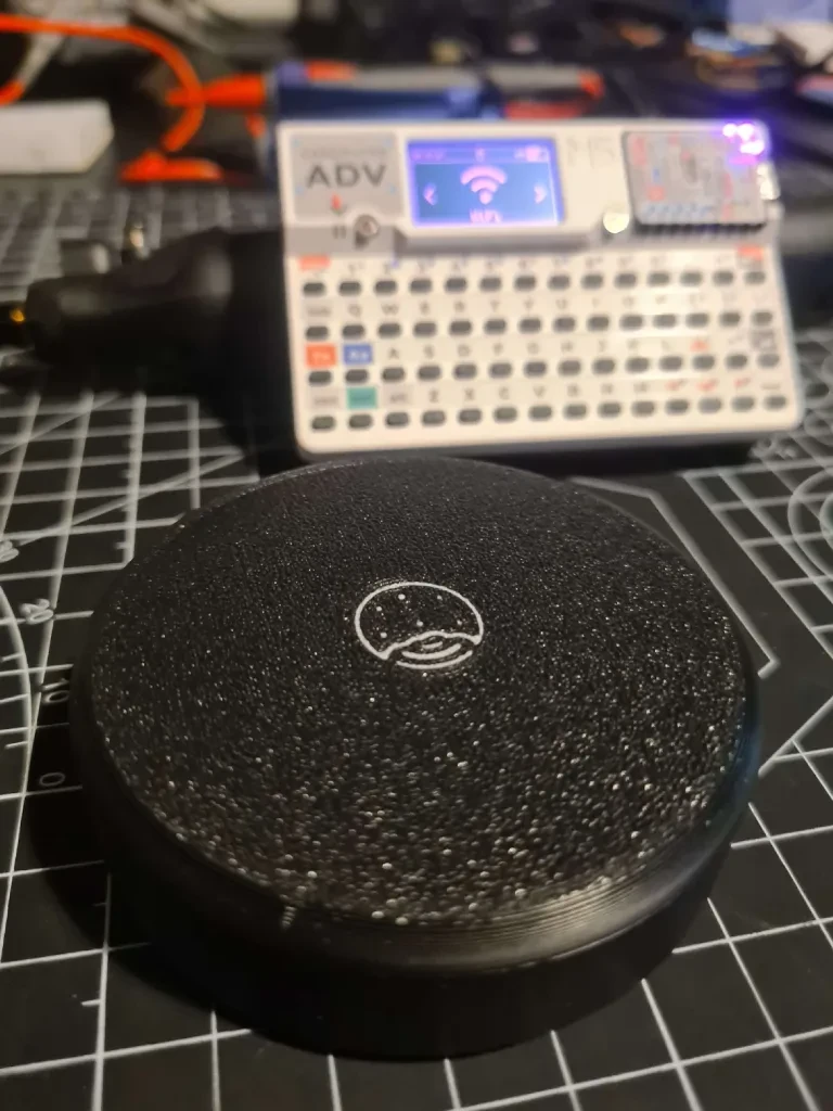 Round black 3D-printed device case on a cutting mat with a Cardputer behind it