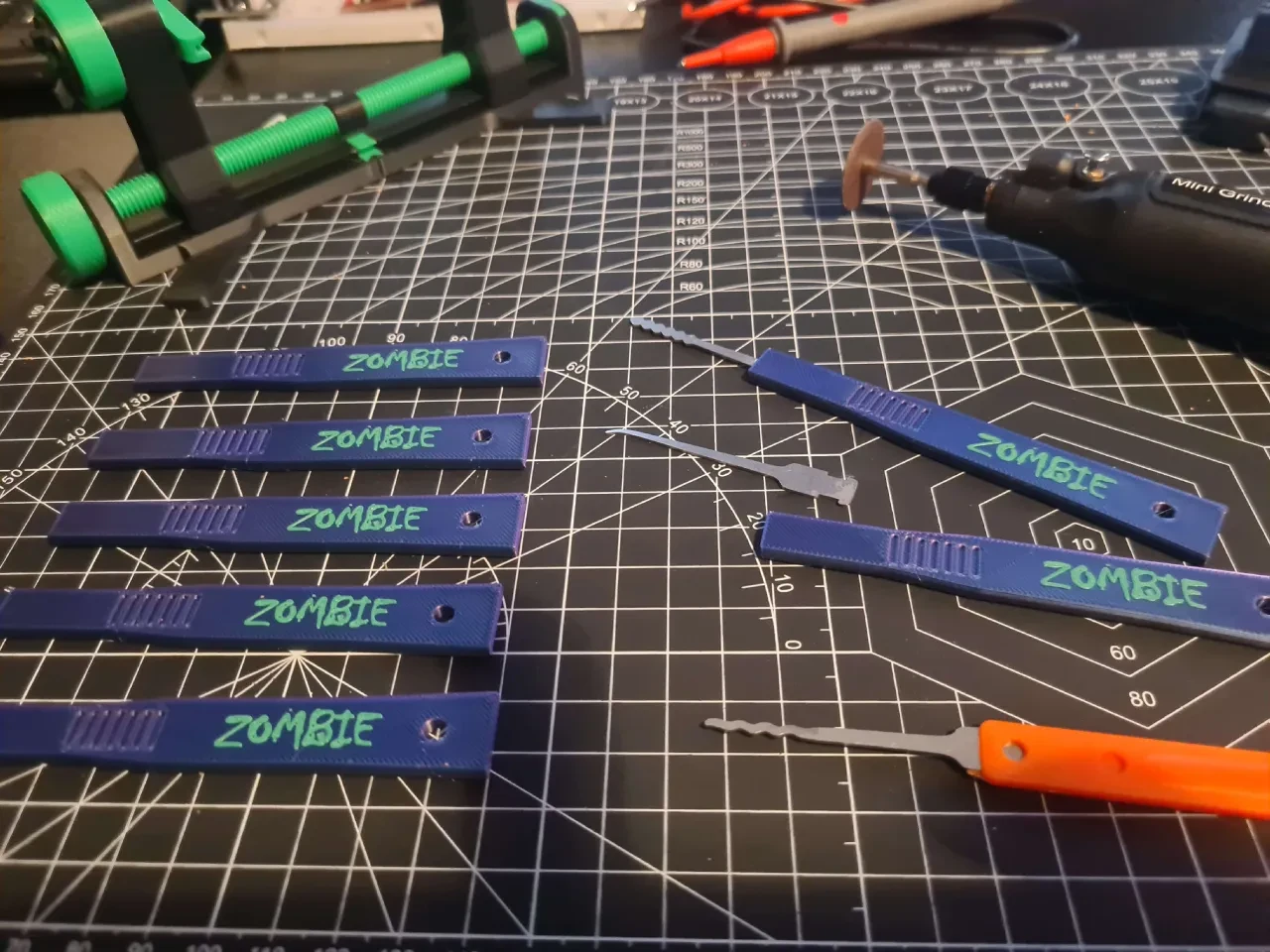 Blue 3D-printed lock-pick handles marked ZOMBIE beside loose picks and a rotary tool on a cutting mat